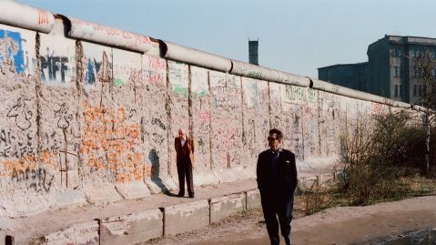 Mann im schwarzen Anzug und Sonnenbrille geht entlang der graffitibedeckten Berliner Mauer, während ein anderer in einem leuchtend Anzug im Hintergrund steht.