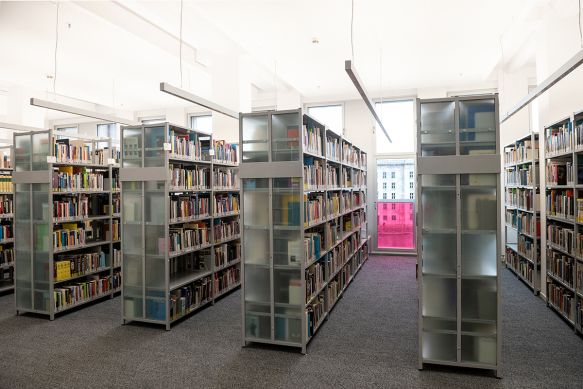 Several shelves with books are located in the library in front of a large window wall.