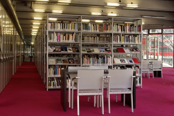 Ein Einblick in die Bibliothek mit Metallregalen, zahlreichen Büchern, Sitzplätzen, einem roten Teppich und einem Ausblick auf das Sony-Center am Potsdamer Platz.