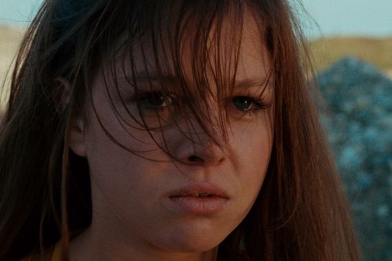 Close-up of a young woman with long hair in the wind, looking serious, with a blurred rocky landscape in warm, natural light.