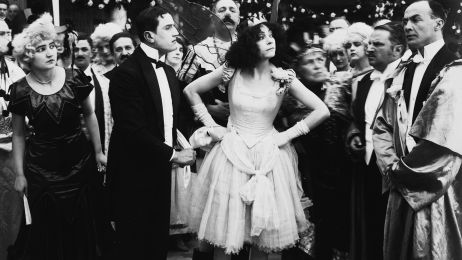 The actress Asta Nielsen stands in a dress and crown with her arms on her hips in a festively dressed group and looks challengingly to the side.