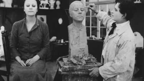 In a studio, a woman sits as a model for a sculptor with her eyes closed.